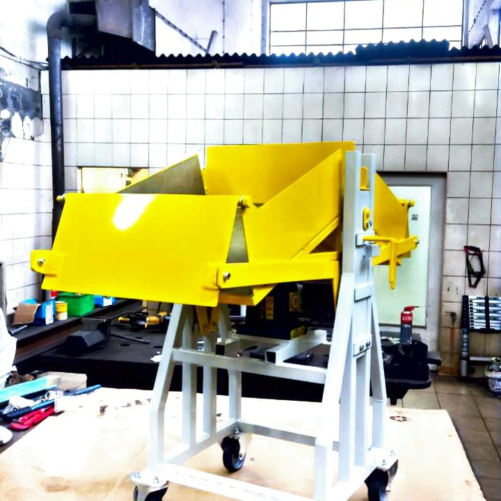 Żółta metalowa konstrukcja na kółkach w warsztacie z białą kafelkową ścianą w tle. Konstrukcja posiada elementy przypominające zsyp lub podajnik.