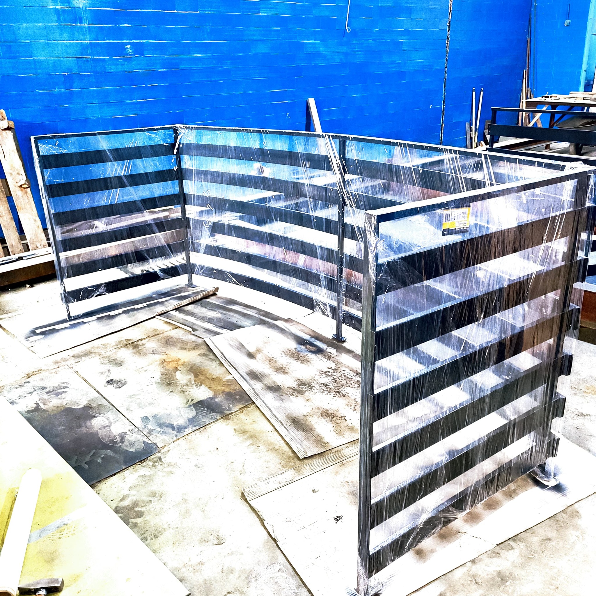 Fragment stalowej konstrukcji z poziomymi poprzeczkami, owiniętej folią ochronną, na tle niebieskiej ściany i fragmentów blachy na podłodze.