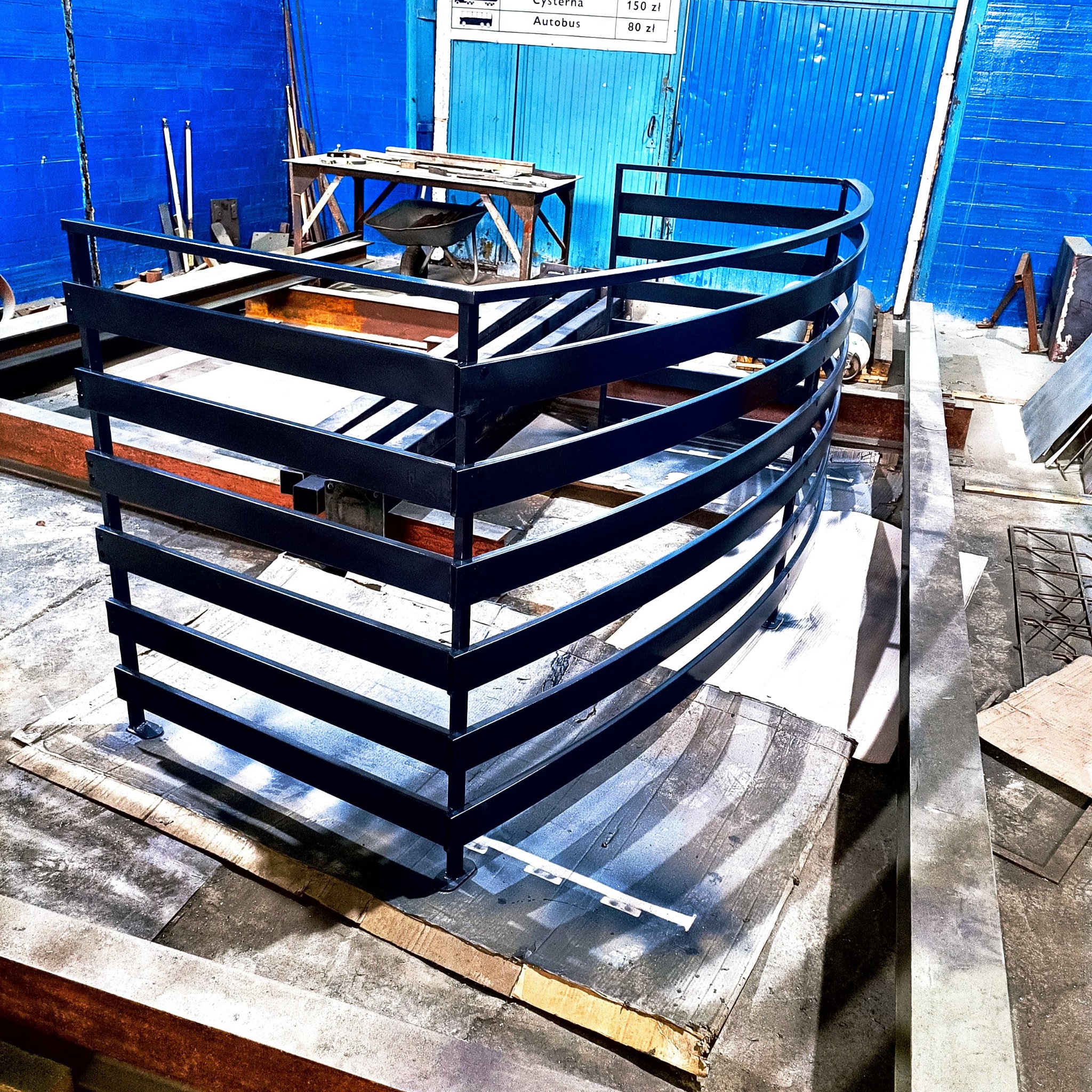 Częściowo zmontowana, czarna, metalowa konstrukcja balustrady o zakrzywionym kształcie, umieszczona w warsztacie na tekturze ochronnej, na tle niebieskiej ściany i warsztatowego stołu.