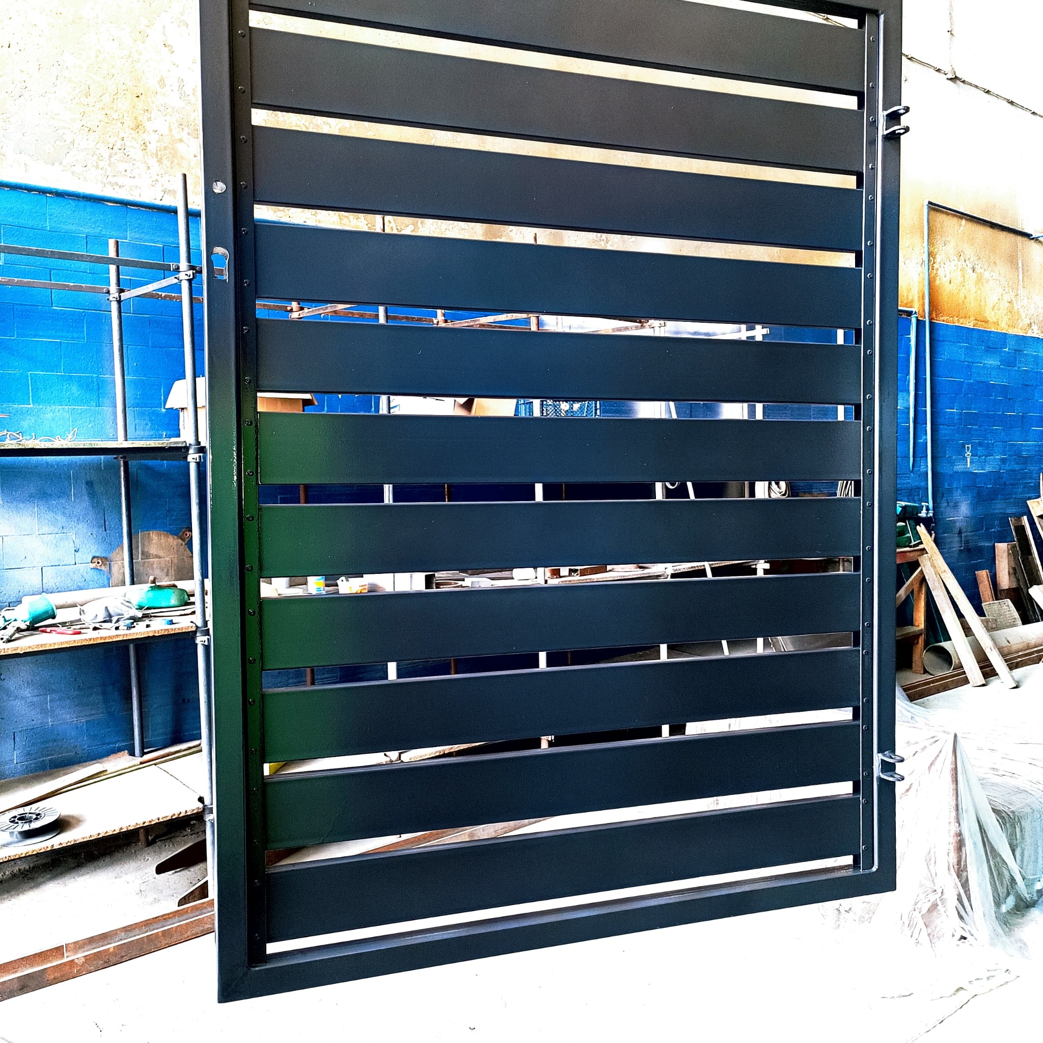 Ciemnoszara brama z poziomych profili, widoczna w warsztacie z niebieską ścianą i rusztowaniem w tle.