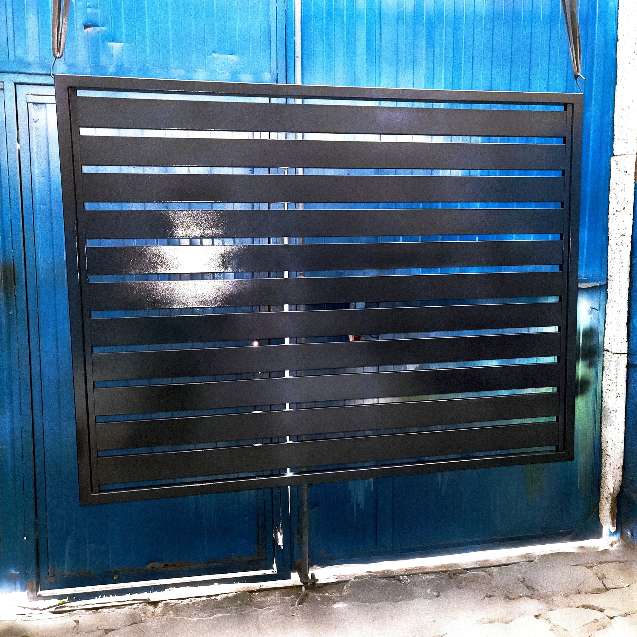 Czarna, pozioma brama lamelowa w trakcie malowania proszkowego, zawieszona na haku, na tle niebieskiej bramy w Nowej Soli.