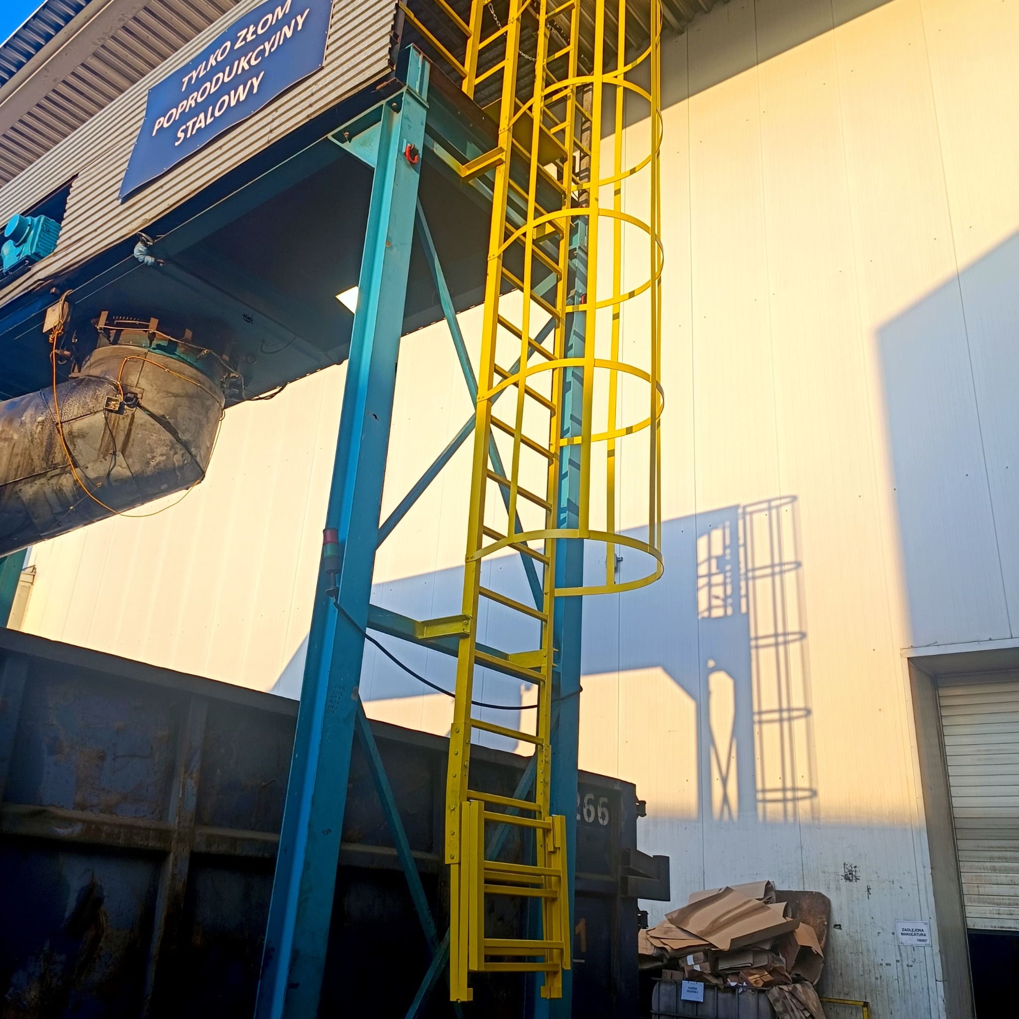 Żółta drabina z zabezpieczeniem przymocowana do niebieskiej konstrukcji stalowej przy hali produkcyjnej z napisem 'Tylko złom poprodukcyjny stalowy'.