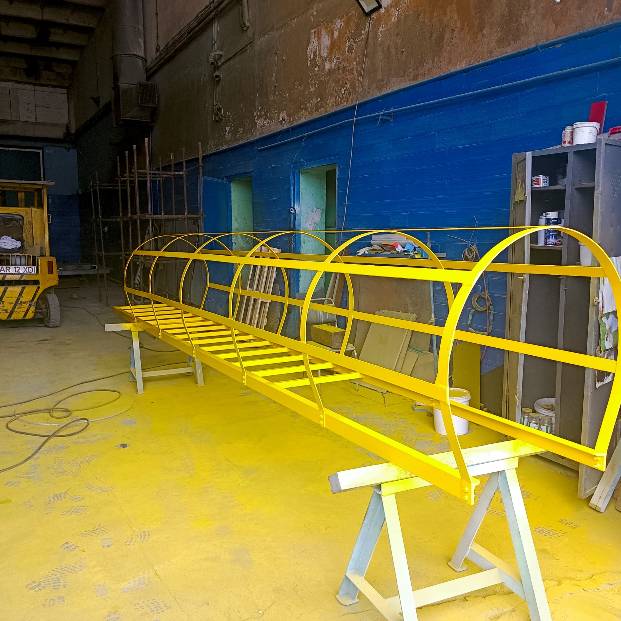 Żółta metalowa konstrukcja schodów z zabezpieczeniem klatkowym, świeżo pomalowana, leżąca na kozłach w warsztacie, na podłodze ślady farby i stóp, w tle widać wózek widłowy.