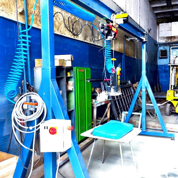 Wnętrze hali produkcyjnej z suwnicą podciśnieniową do przenoszenia elementów; widoczne niebieskie ramy konstrukcyjne, biała skrzynka sterownicza z czerwonym przyciskiem, zielona szafka, niebieskie...