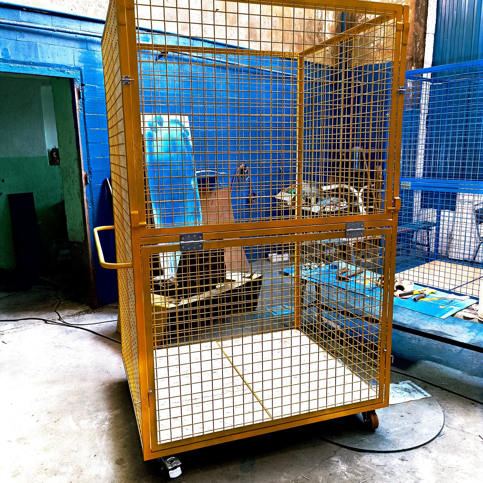 Żółta, metalowa klatka z siatki na kółkach, z otwartymi drzwiami, stojąca w warsztacie z niebieskimi ścianami i podłogą, widoczne narzędzia w środku i na zewnątrz klatki.