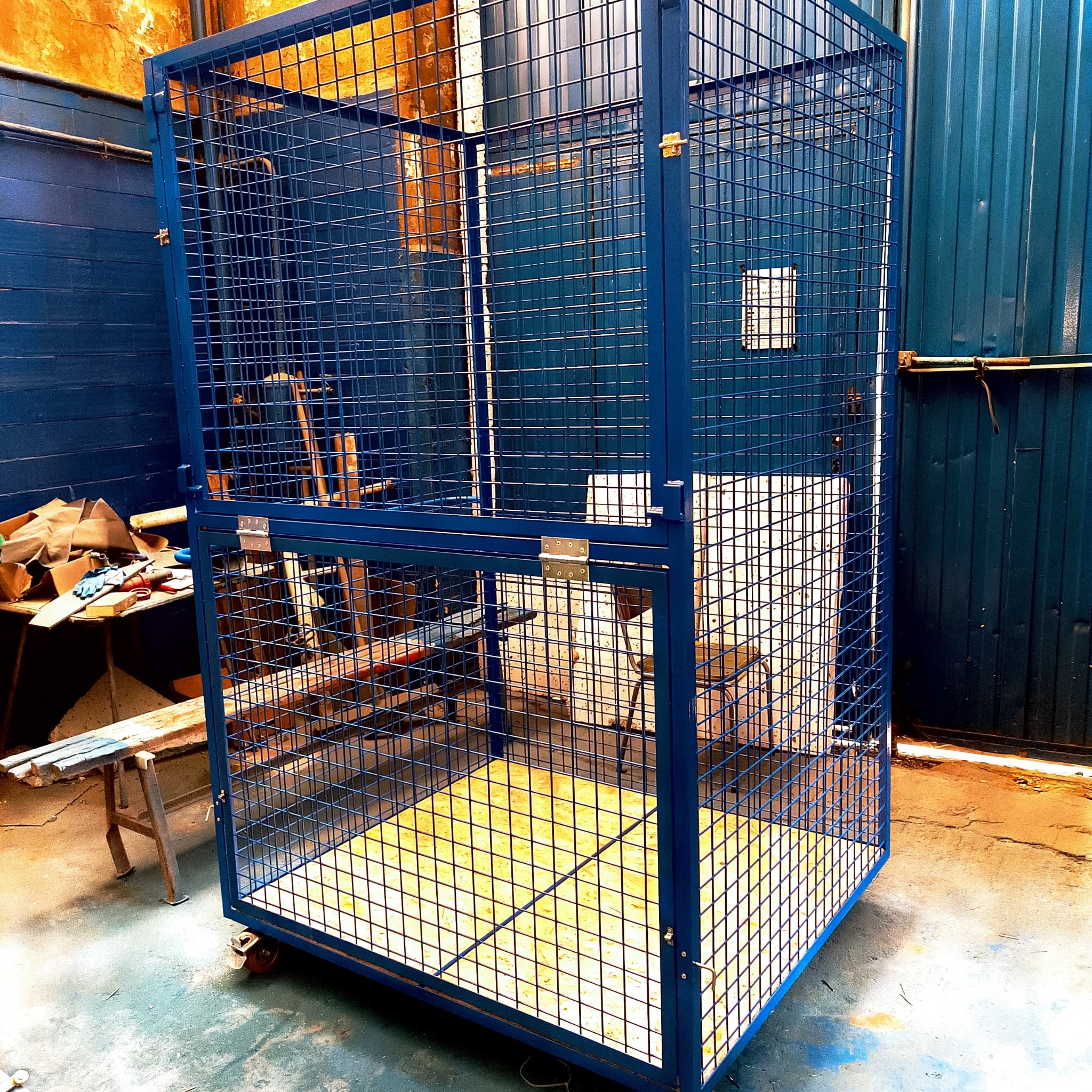 Niebieska, metalowa klatka magazynowa z kratką, na kółkach, z żółtą podłogą, widoczna w warsztacie, obok stół z narzędziami.