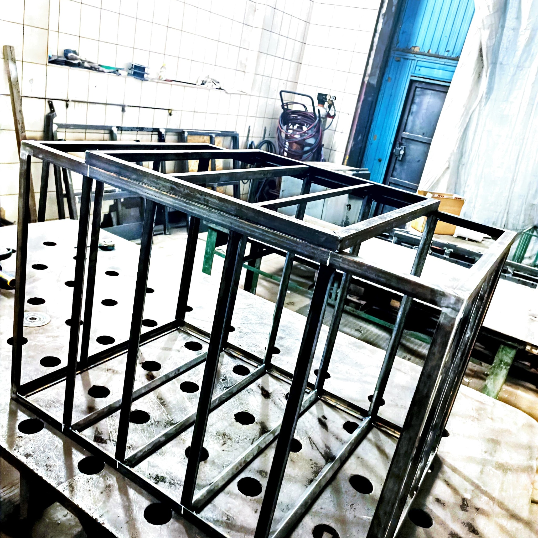 Surowa, spawana konstrukcja stalowa w kształcie klatki na perforowanym stole warsztatowym w hali produkcyjnej, widoczne ślady spawania i niedoskonałości materiału.