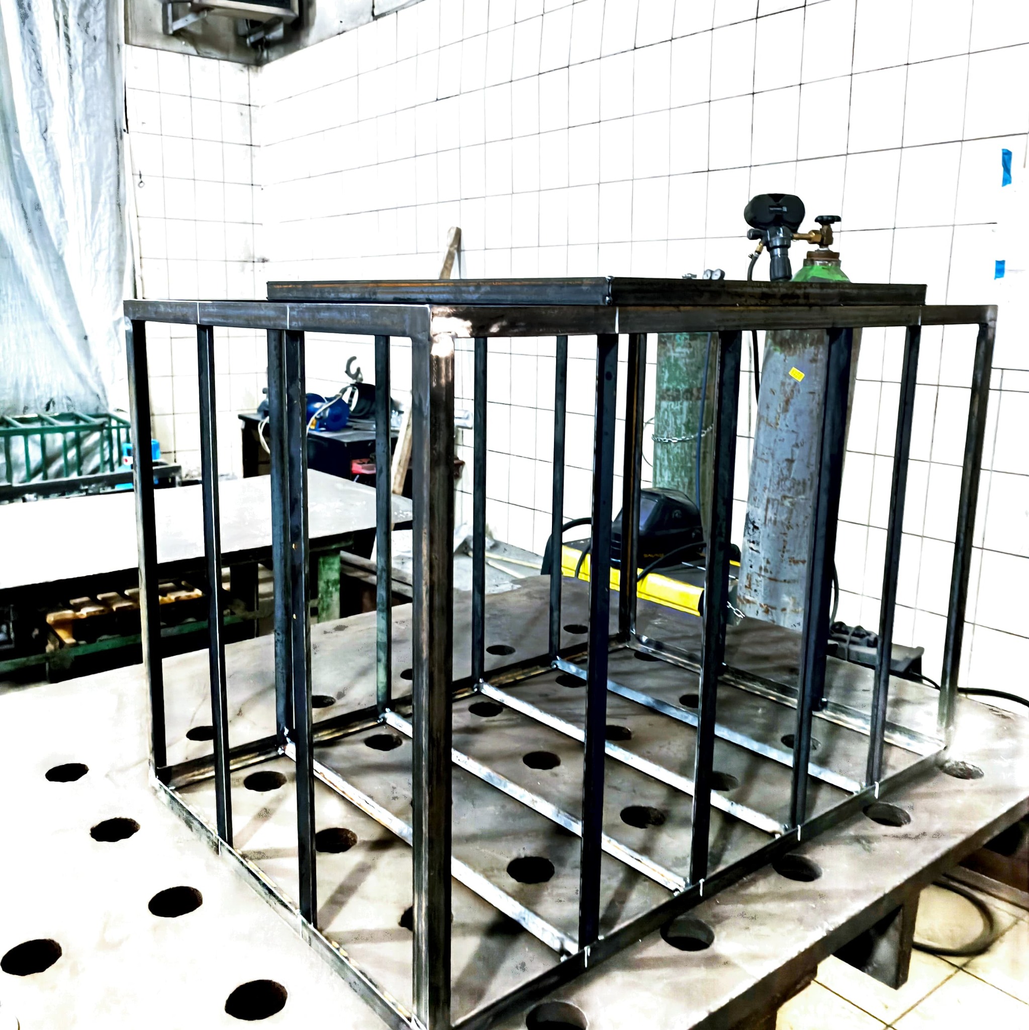 Konstrukcja stalowa w trakcie spawania na stole spawalniczym z otworami, widoczne butle z gazem i sprzęt spawalniczy w tle, hala produkcyjna z białymi kafelkami na ścianach.