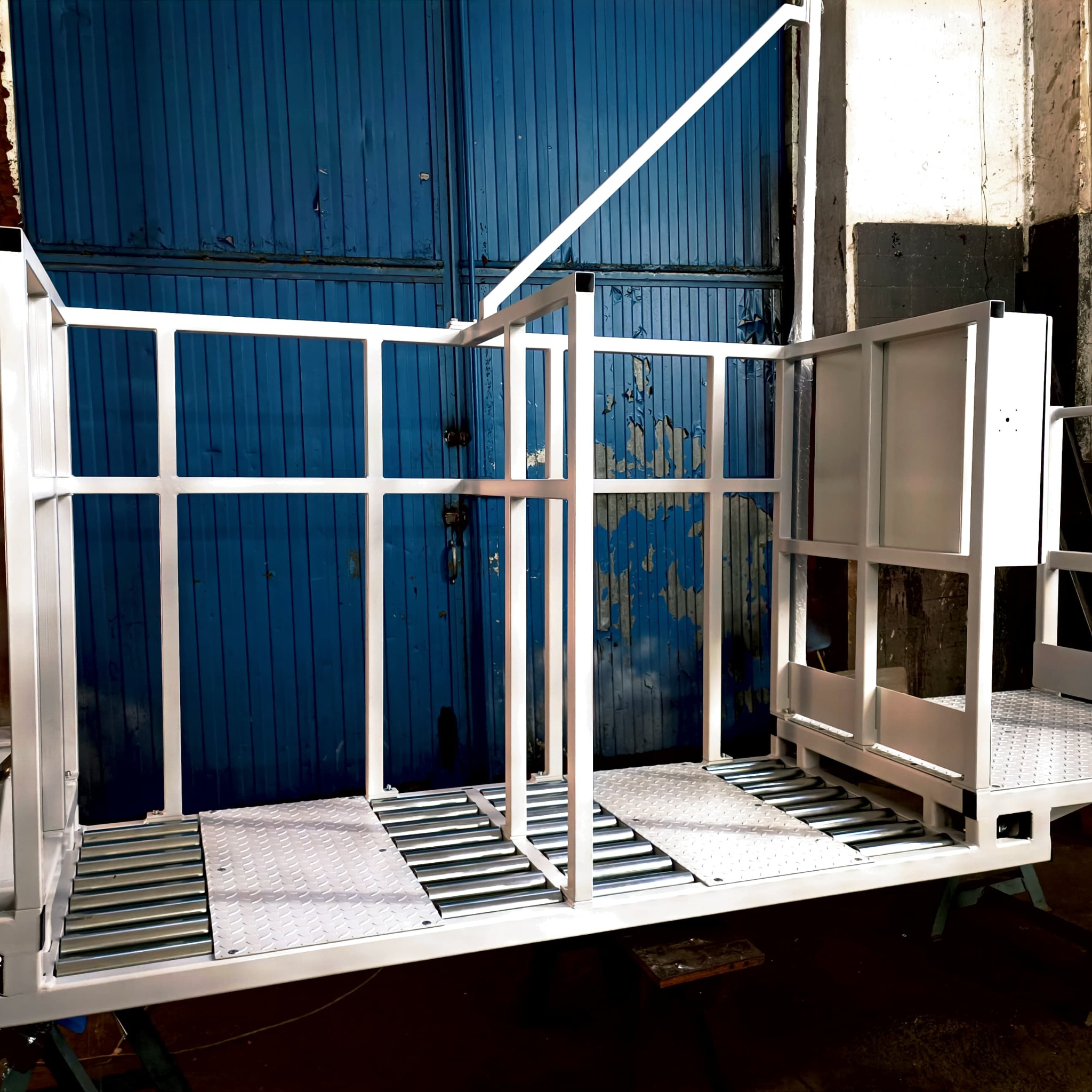 Surowa, metalowa konstrukcja wózka platformowego z białymi, kwadratowymi profilami i podłogą z blachy ryflowanej na tle niebieskiej, łuszczącej się bramy garażowej.