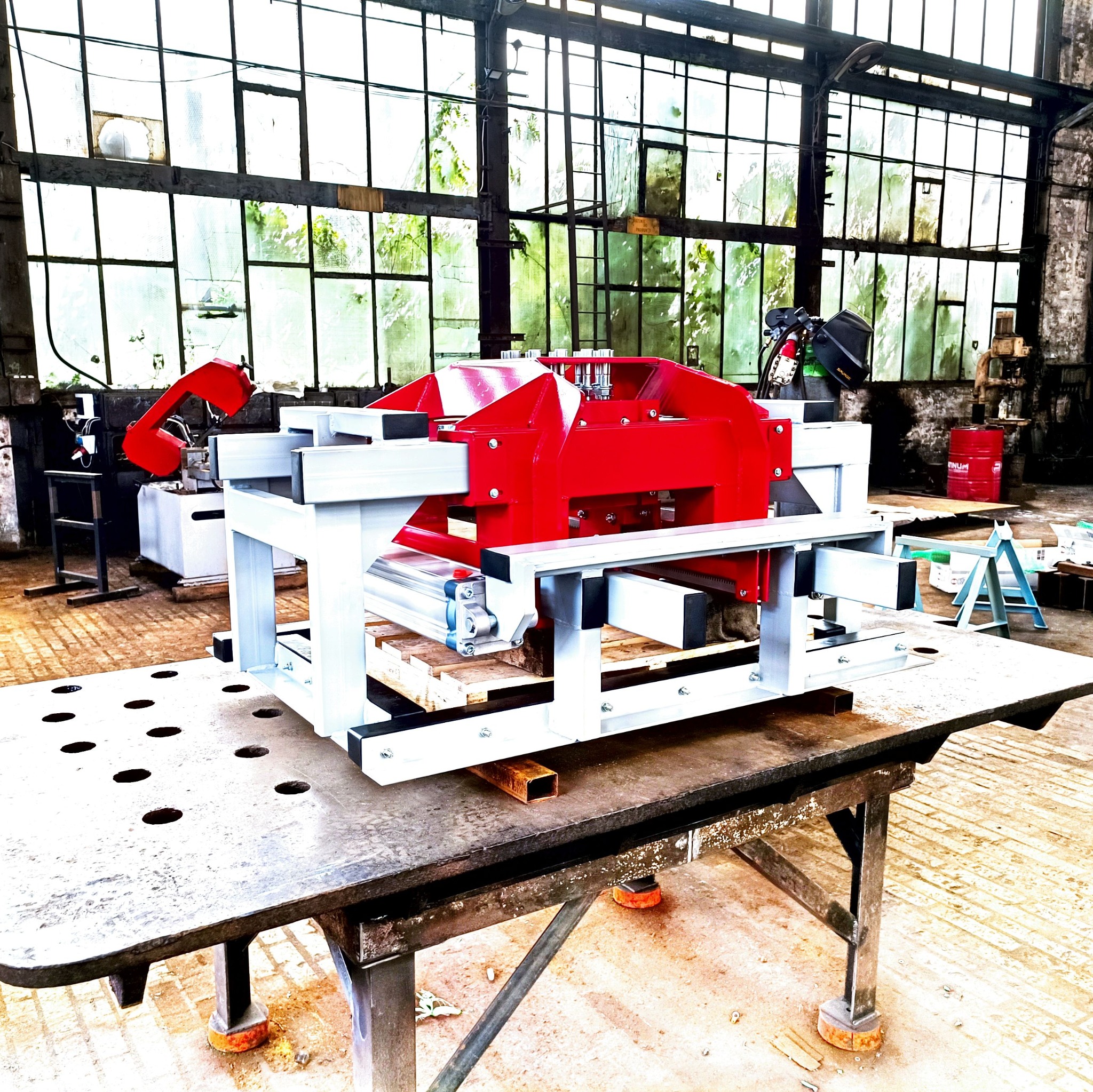 Czerwono-biała konstrukcja stalowa na stole spawalniczym z otworami, w tle okna hali przemysłowej i sprzęt spawalniczy.