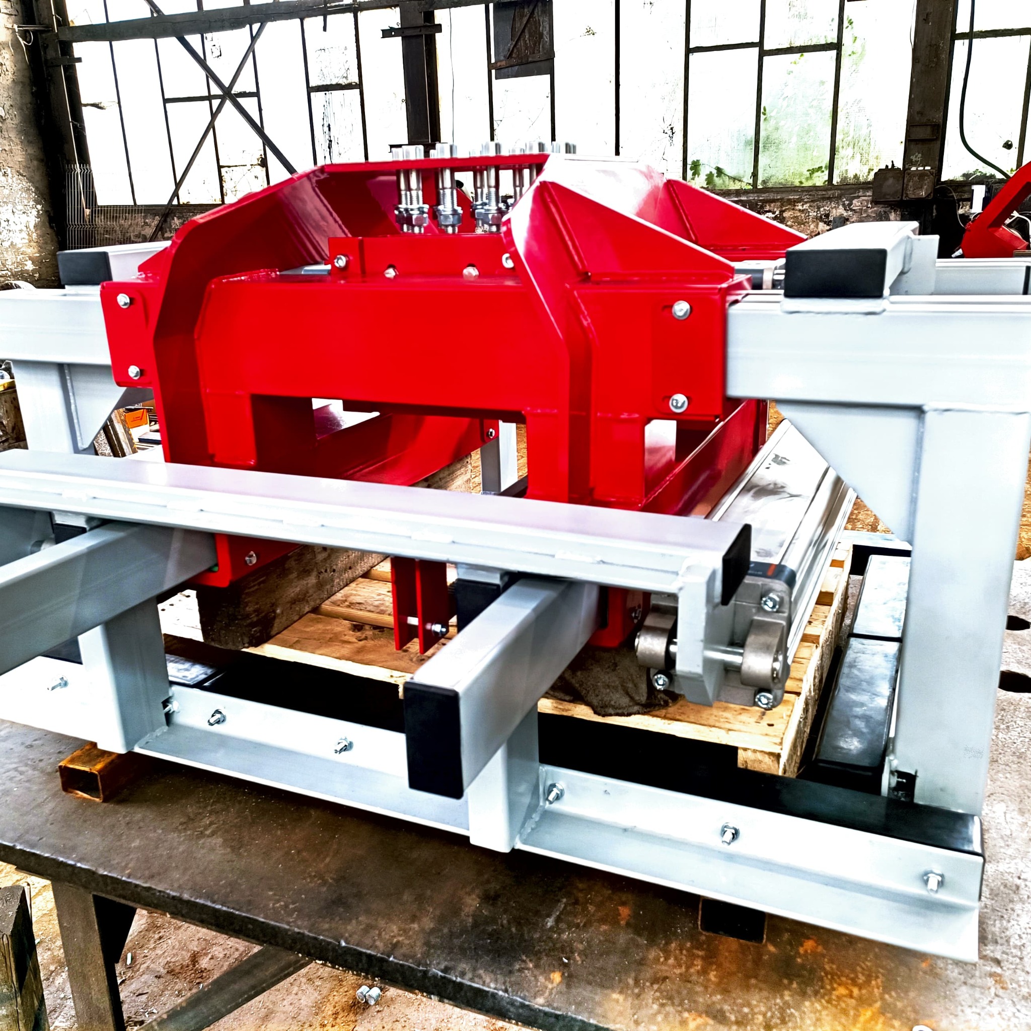 Czerwono-szara maszyna do obróbki metalu, z widocznymi śrubami i siłownikami, umieszczona na palecie w hali produkcyjnej.