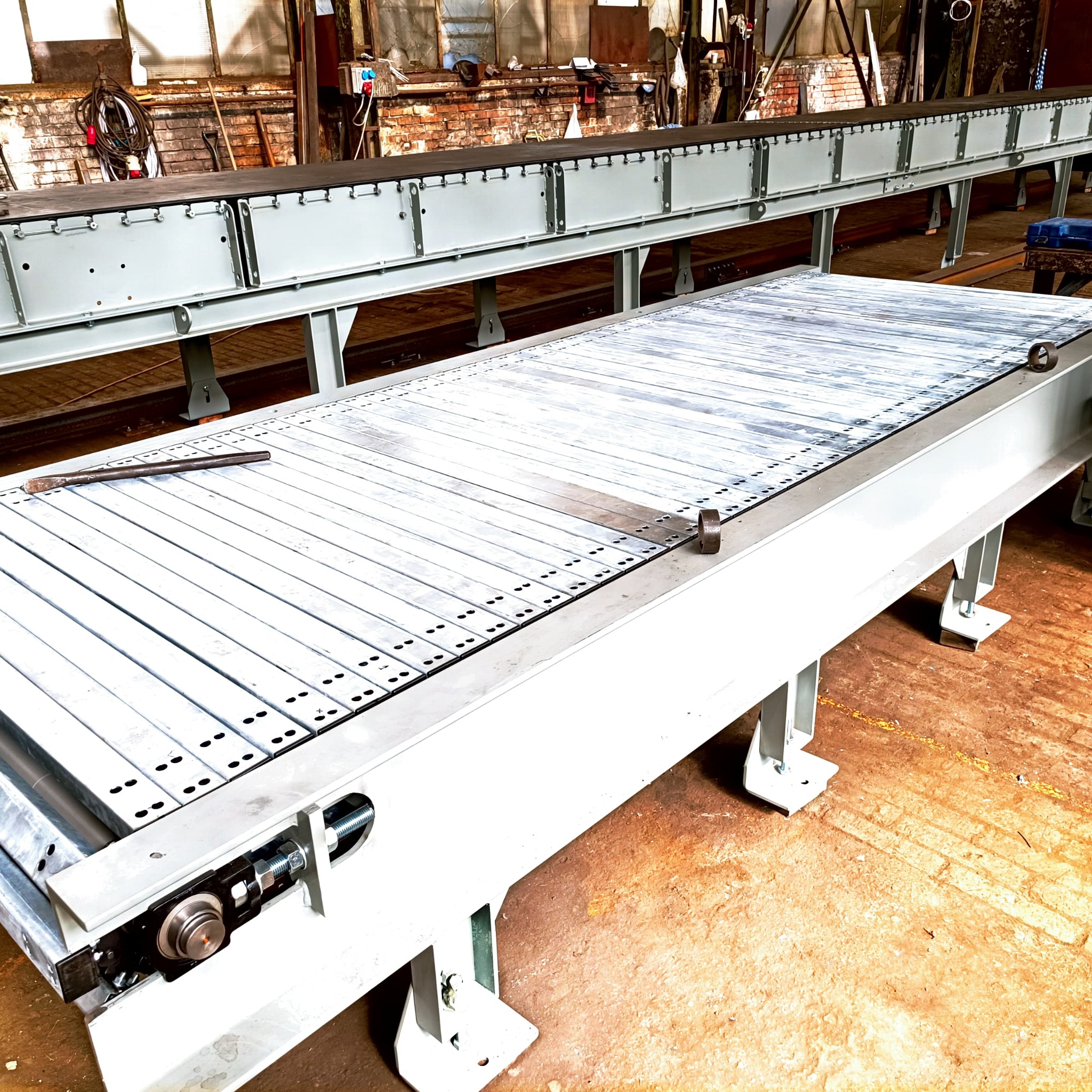 Długa, metalowa konstrukcja przenośnika taśmowego z elementami łożyskowymi, widoczna w hali produkcyjnej z ceglaną ścianą w tle.