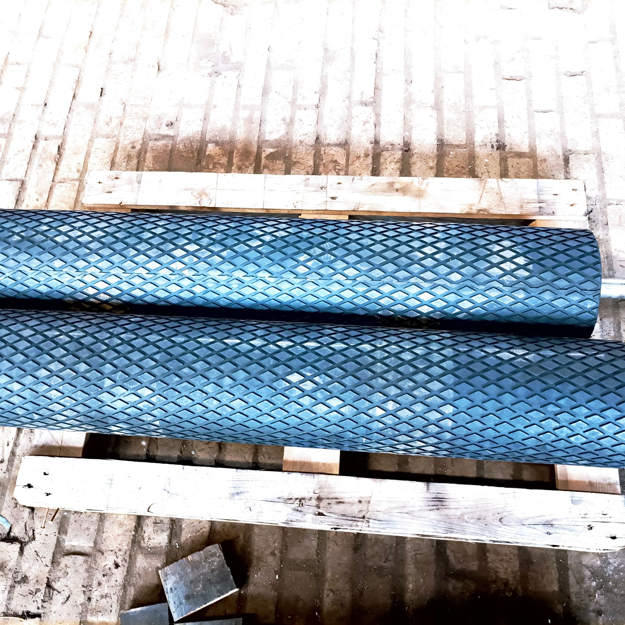 Dwa niebieskie, gumowane wałki z diamentowym wzorem, ułożone na drewnianej palecie w magazynie.