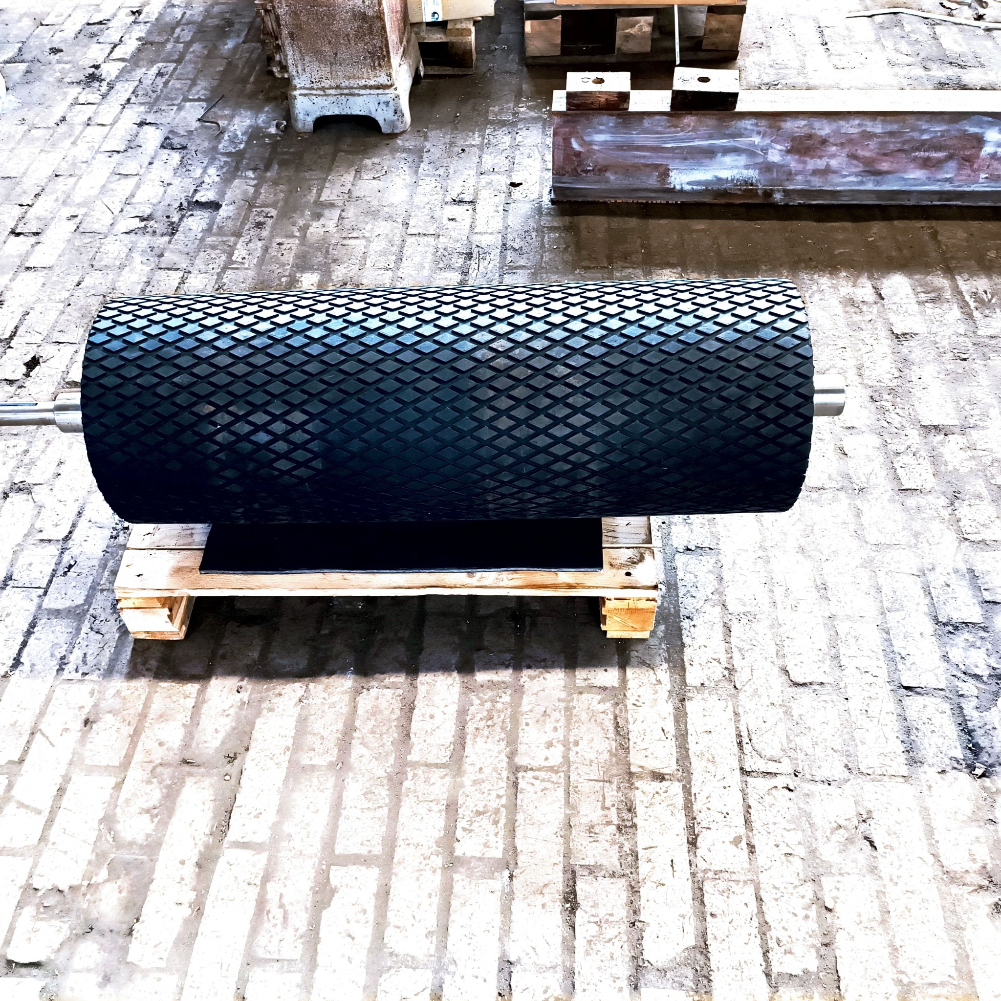 Czarny, gumowany walec z romboidalnym wzorem powierzchni, umieszczony na drewnianej palecie, widoczny na tle betonowej posadzki w hali produkcyjnej.