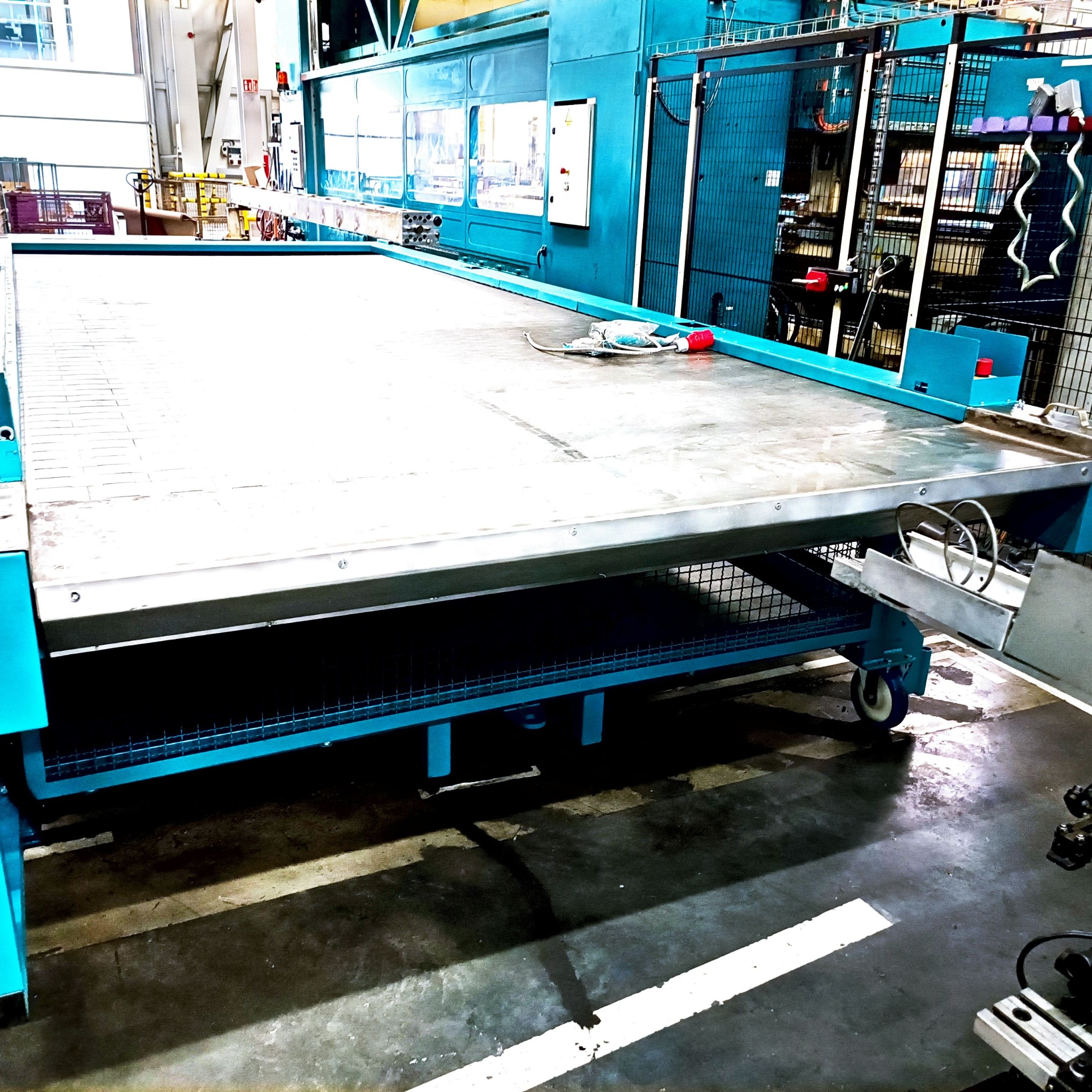 Duży, turkusowy stół roboczy z metalową siatką pod spodem i narzędziami na blacie, widziany w hali przemysłowej.