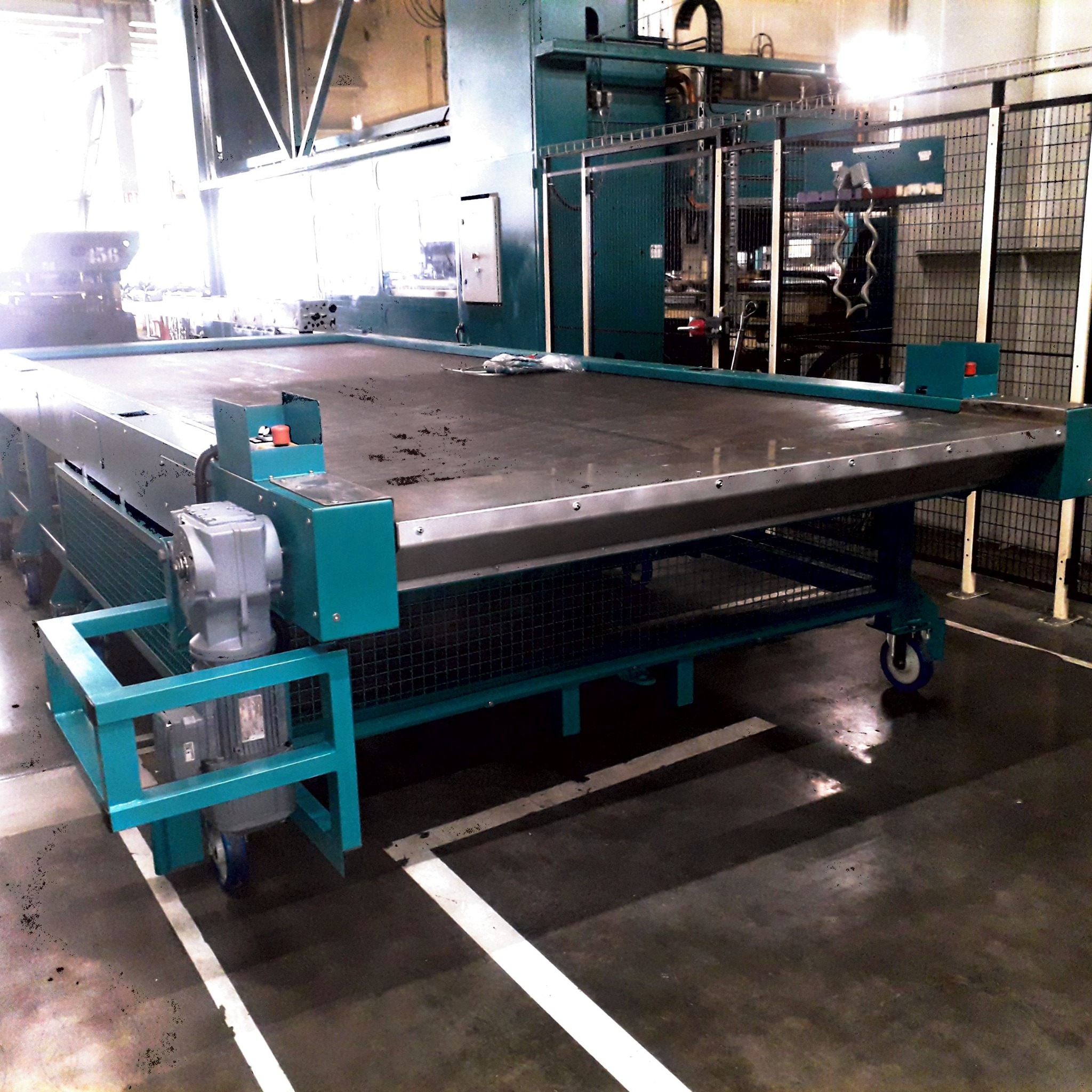 Duży, turkusowy stół roboczy z metalową konstrukcją i kółkami, stojący w hali produkcyjnej, z widocznym w tle fragmentem maszyny przemysłowej i elementami zabezpieczeń.