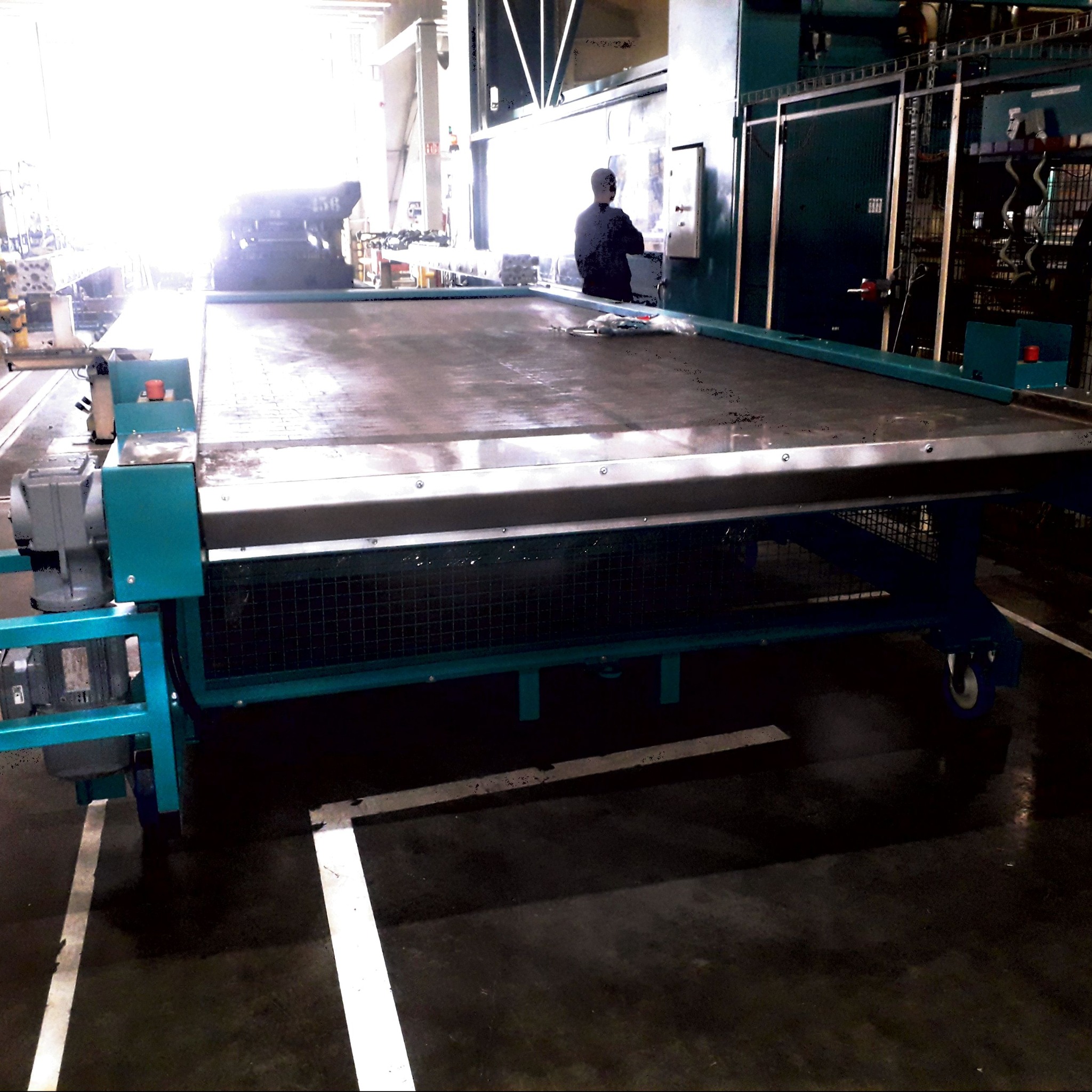 Duży, mobilny stół roboczy z metalowym blatem i turkusową konstrukcją nośną na kółkach, widoczny w hali produkcyjnej z operatorem przy panelu sterowania w tle.