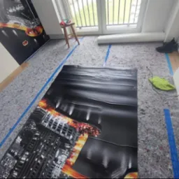 Przygotowanie podłoża pod panele z folią i izolacją. Widoczne taśmy wyznaczające obszar roboczy i częściowo położona fototapeta na ścianie.