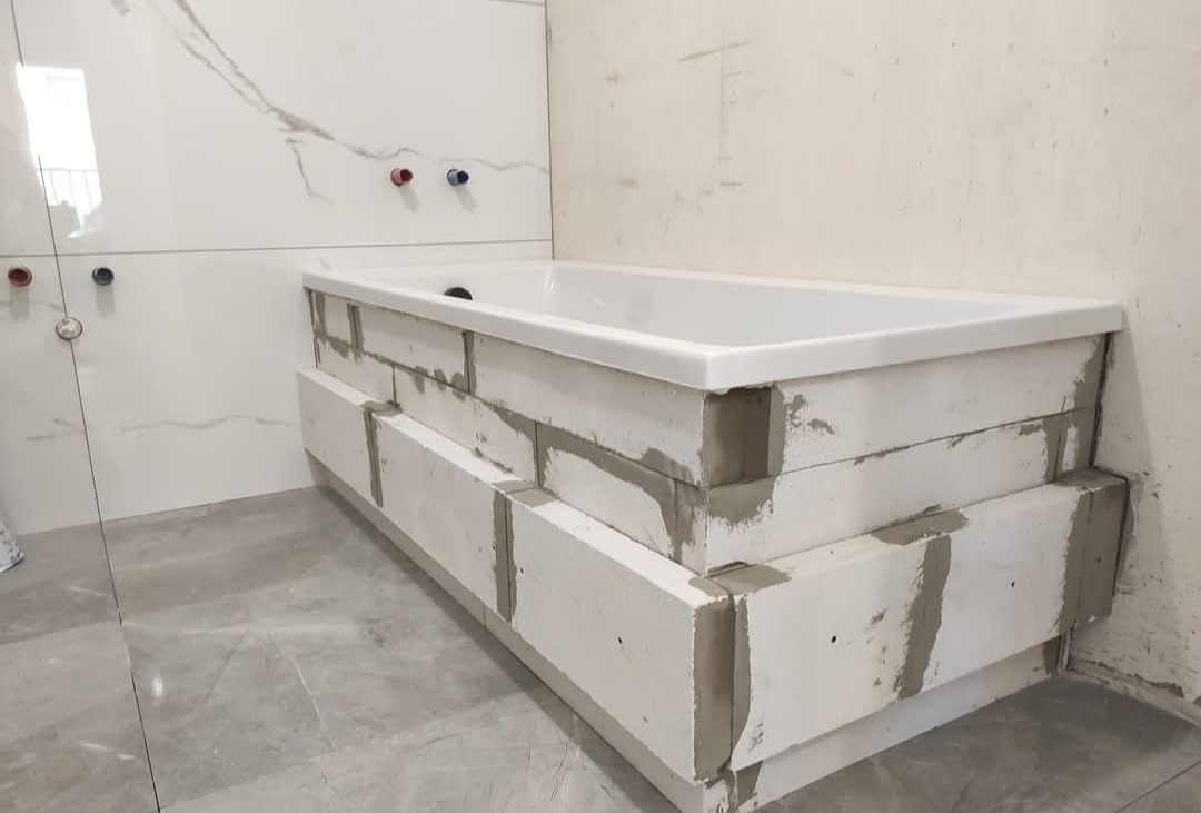 Biała wanna obudowana bloczkami z betonu komórkowego w trakcie prac wykończeniowych łazienki. Widoczne przyłącza wodne na ścianie z płytkami imitującymi marmur.