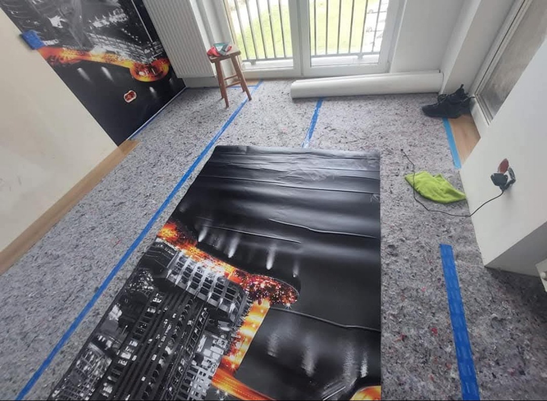 Przygotowanie podłoża pod panele z folią i izolacją. Widoczne taśmy wyznaczające obszar roboczy i częściowo położona fototapeta na ścianie.