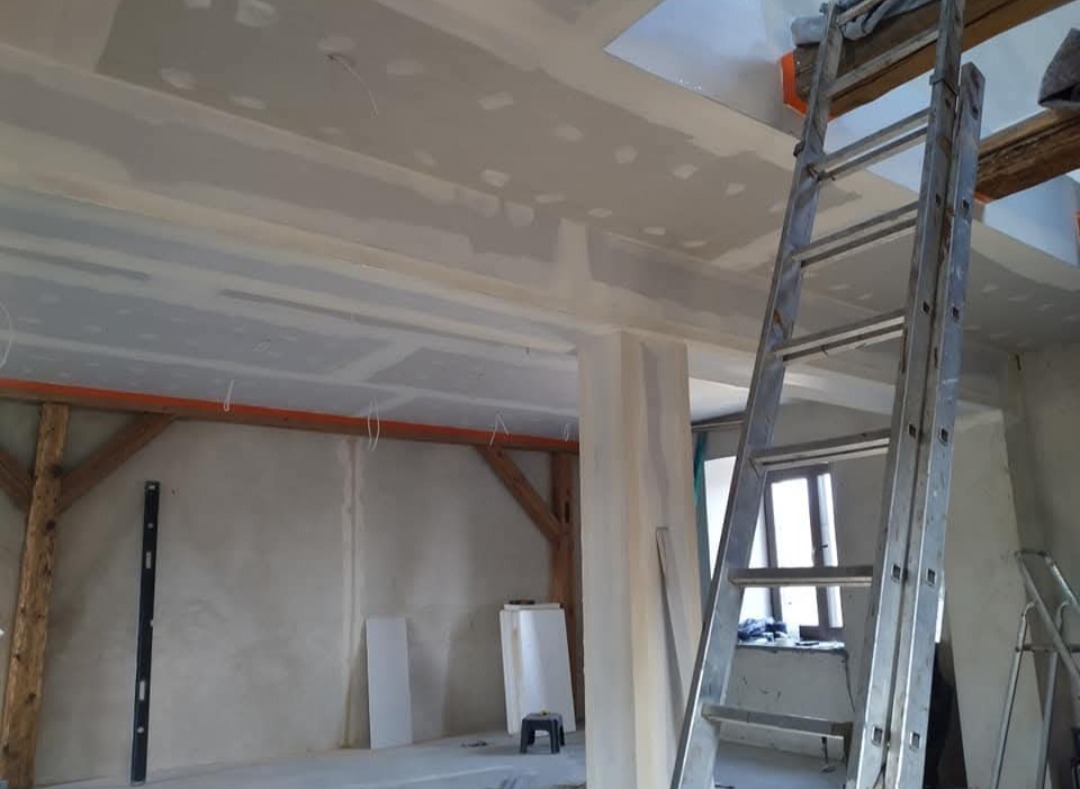Wnętrze w trakcie prac wykończeniowych: zamontowane płyty gipsowe na suficie, widoczna drabina, drewniane elementy konstrukcyjne i poziomica na ścianie.