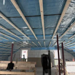 Konstrukcja drewniana poddasza z folią w kolorze niebieskim, widoczne elementy konstrukcyjne i przygotowane materiały budowlane