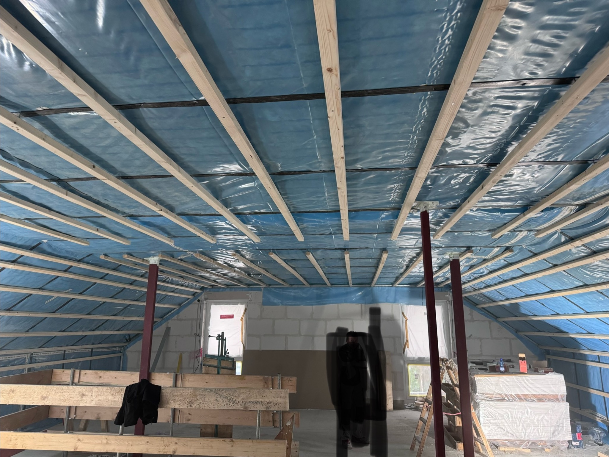 Konstrukcja drewniana poddasza z folią w kolorze niebieskim, widoczne elementy konstrukcyjne i przygotowane materiały budowlane
