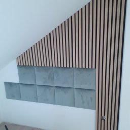Kris - Panel dekoracyjny z drewnianych listew i szarych kwadratowych paneli tapicerowanych na skośnej ścianie poddasza, widoczne elementy montażowe u dołu.