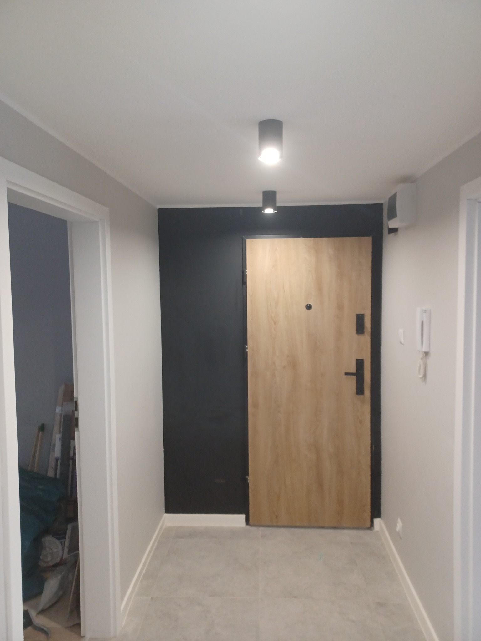 Wnętrze mieszkania po remoncie: Zamontowane drzwi wejściowe z okleiną drewnopodobną na tle czarnej ściany, widoczne oświetlenie punktowe i fragment korytarza z szarą podłogą.