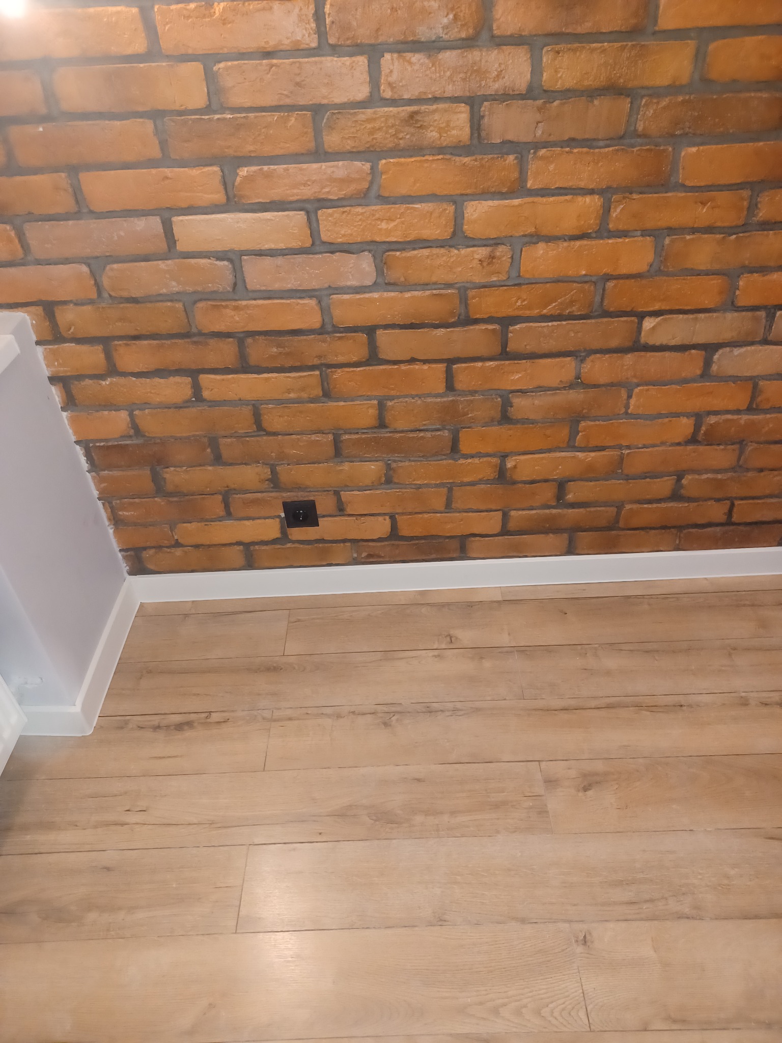 Ściana z cegły dekoracyjnej w odcieniach pomarańczy, z widocznym gniazdkiem elektrycznym i fragmentem jasnej podłogi laminowanej oraz białą listwą przypodłogową.