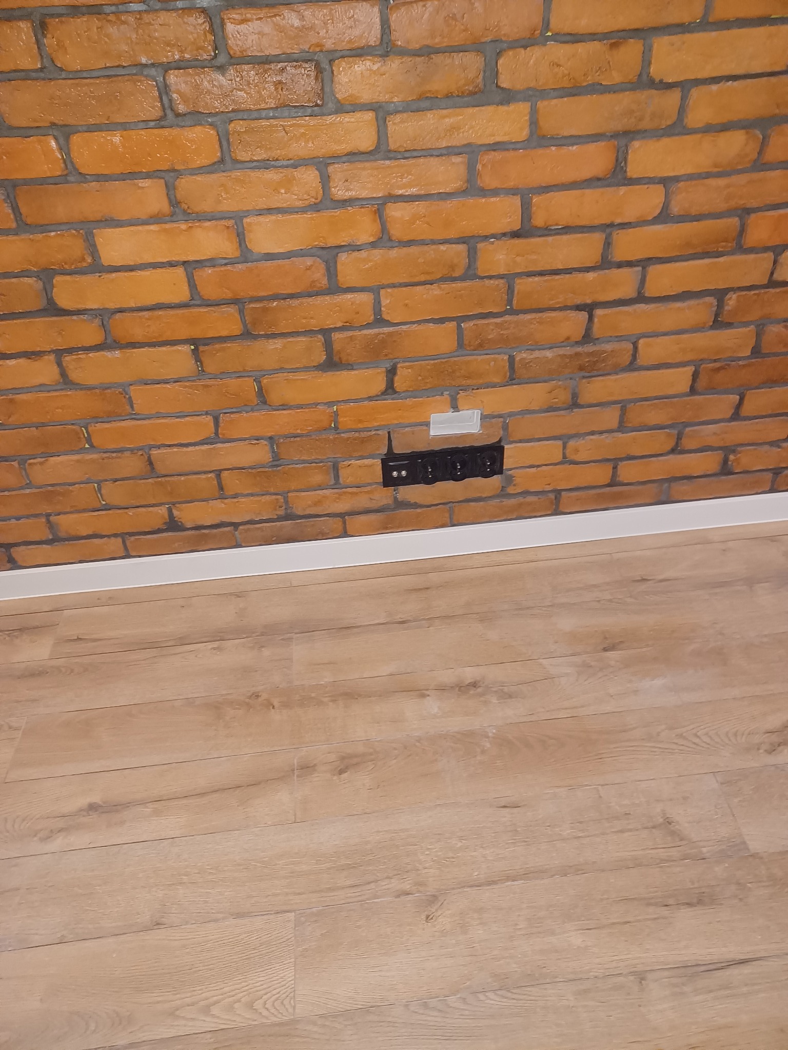 Ściana z cegły dekoracyjnej w odcieniach pomarańczy z widocznym gniazdkiem elektrycznym i listwą przypodłogową, na podłodze panele imitujące drewno.