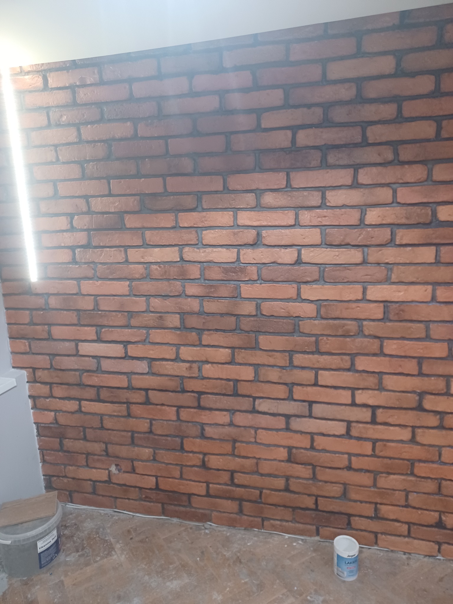 Ściana wykończona cegłą dekoracyjną w odcieniach czerwieni i brązu, z widocznymi fugami, stojące na podłodze wiadro z zaprawą i puszka farby, oświetlona z lewej strony.
