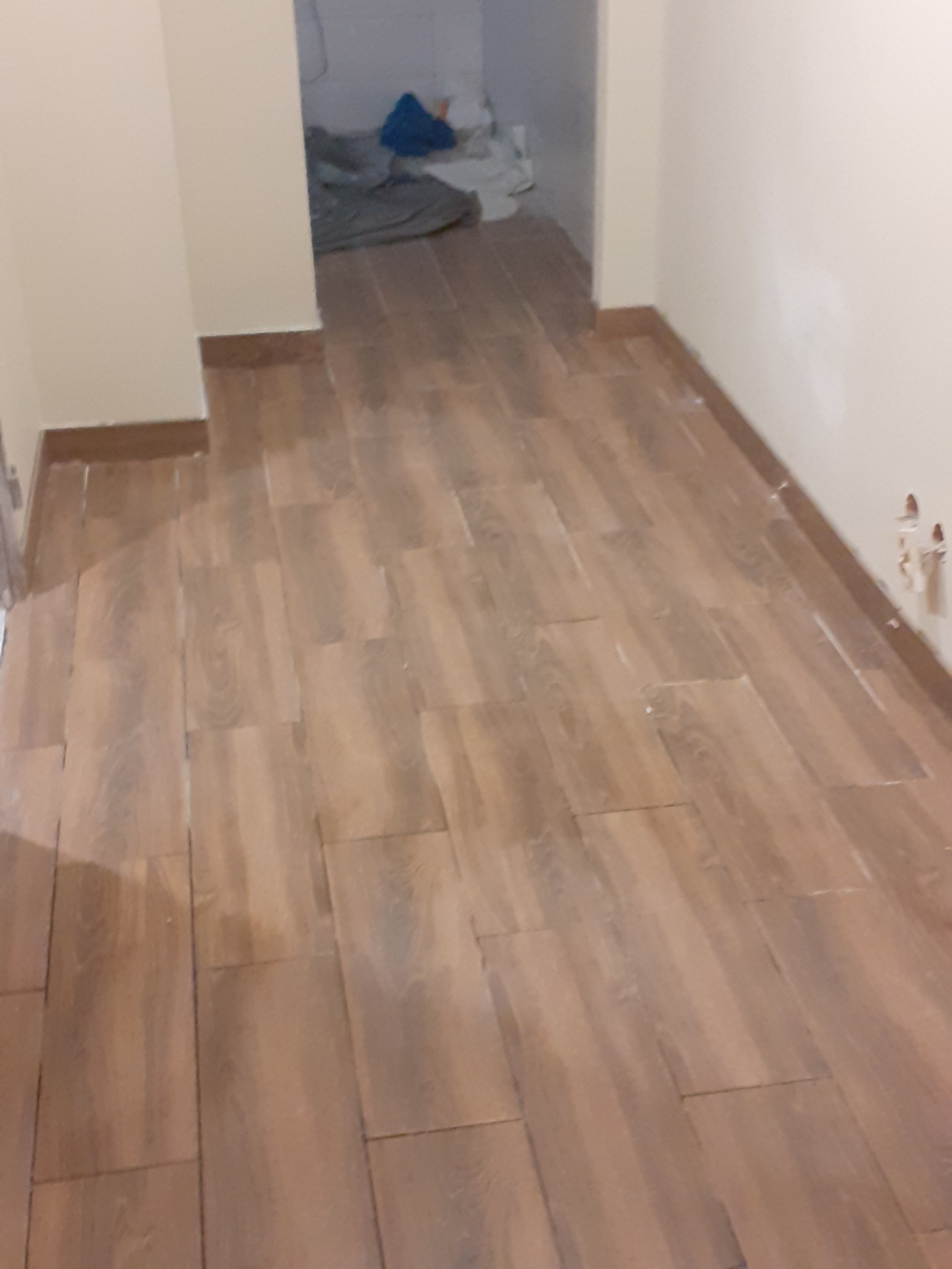 Ułożona, brązowa podłoga z płytek imitujących drewno w trakcie prac wykończeniowych, widoczne niedoskonałości przy ścianach i wystające kable elektryczne.