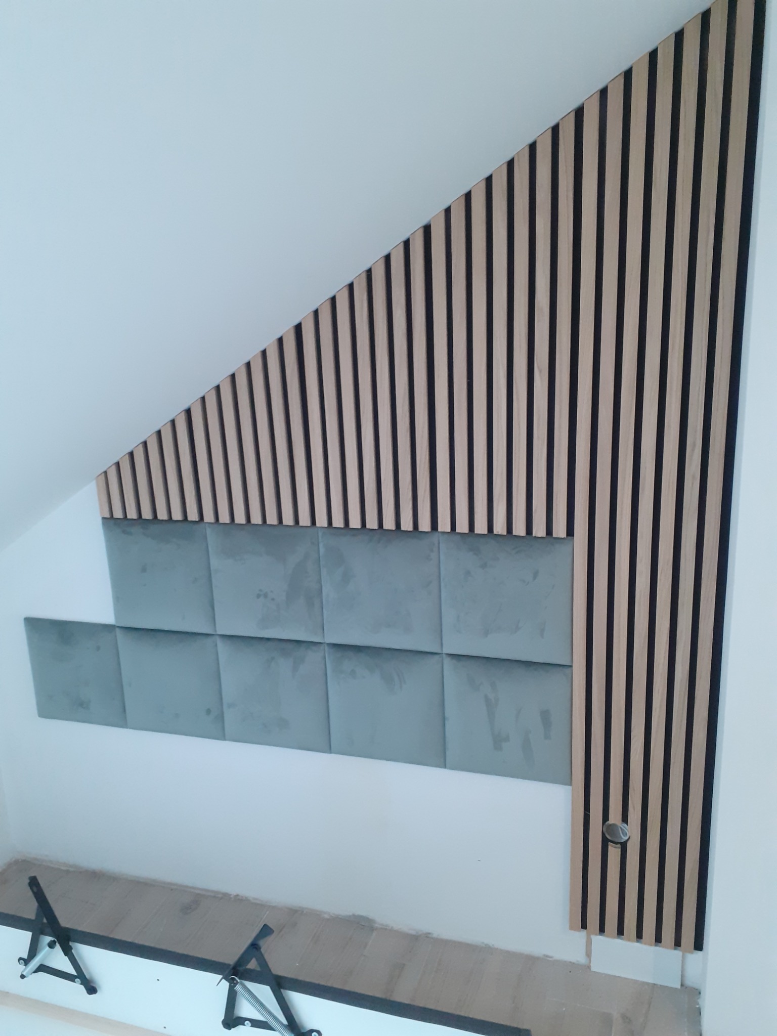 Panel dekoracyjny z drewnianych listew i szarych kwadratowych paneli tapicerowanych na skośnej ścianie poddasza, widoczne elementy montażowe u dołu.