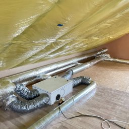 MS-systems - Rekuperacja na poddaszu: centrala wentylacyjna z podłączonymi przewodami wentylacyjnymi, widoczne zabezpieczenie folią paroizolacyjną na skośnym dachu.