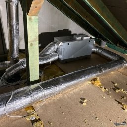 MS-systems - Systemair rekuperator SAVE z podłączonymi przewodami wentylacyjnymi na poddaszu, widoczne elementy konstrukcji dachu i izolacji.