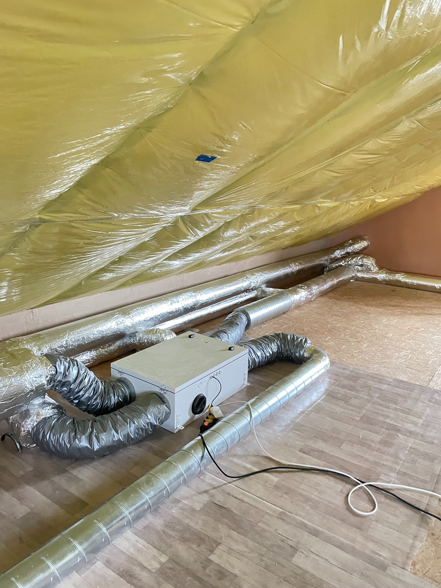 Rekuperacja na poddaszu: centrala wentylacyjna z podłączonymi przewodami wentylacyjnymi, widoczne zabezpieczenie folią paroizolacyjną na skośnym dachu.