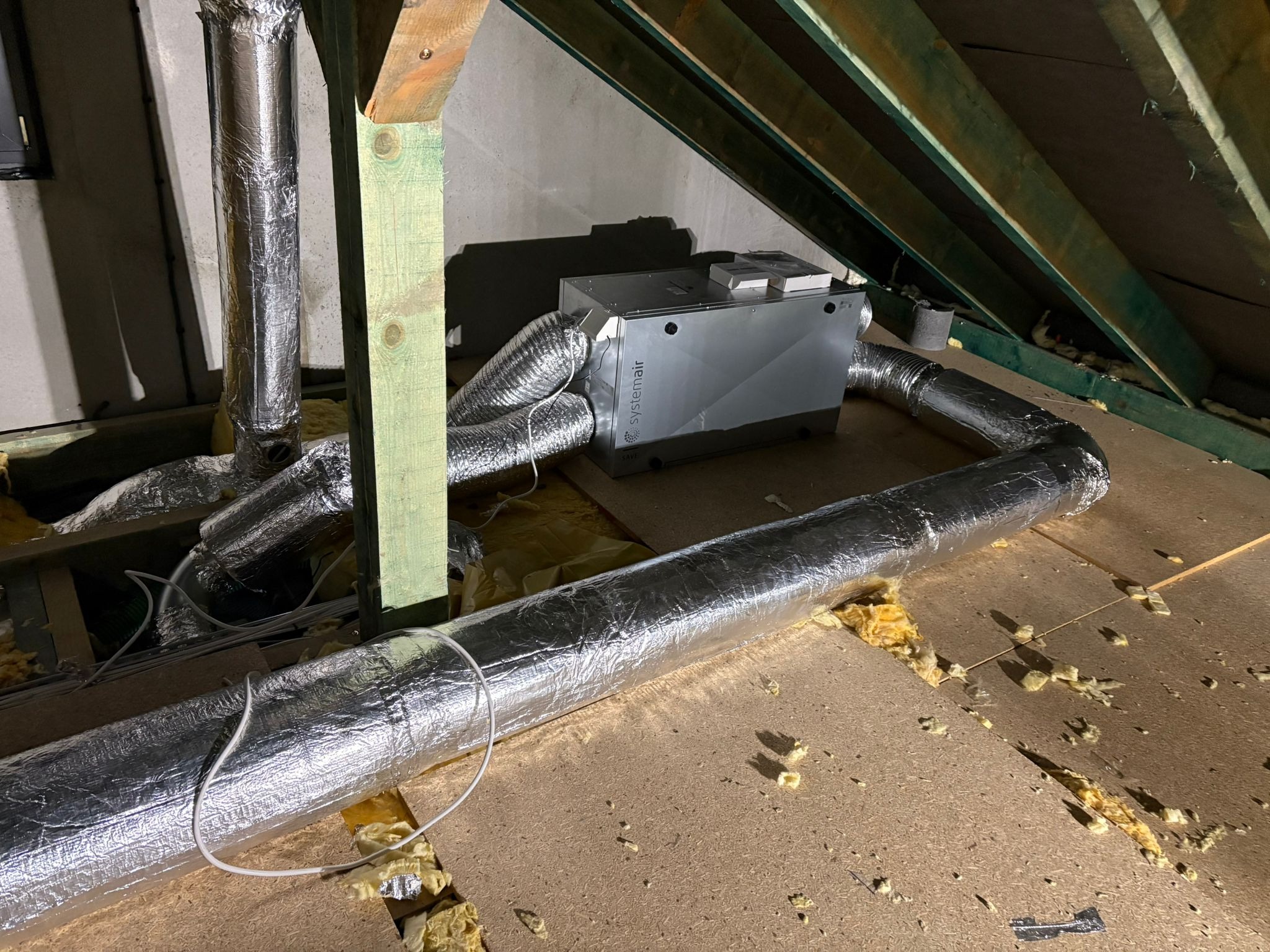 Systemair rekuperator SAVE z podłączonymi przewodami wentylacyjnymi na poddaszu, widoczne elementy konstrukcji dachu i izolacji.