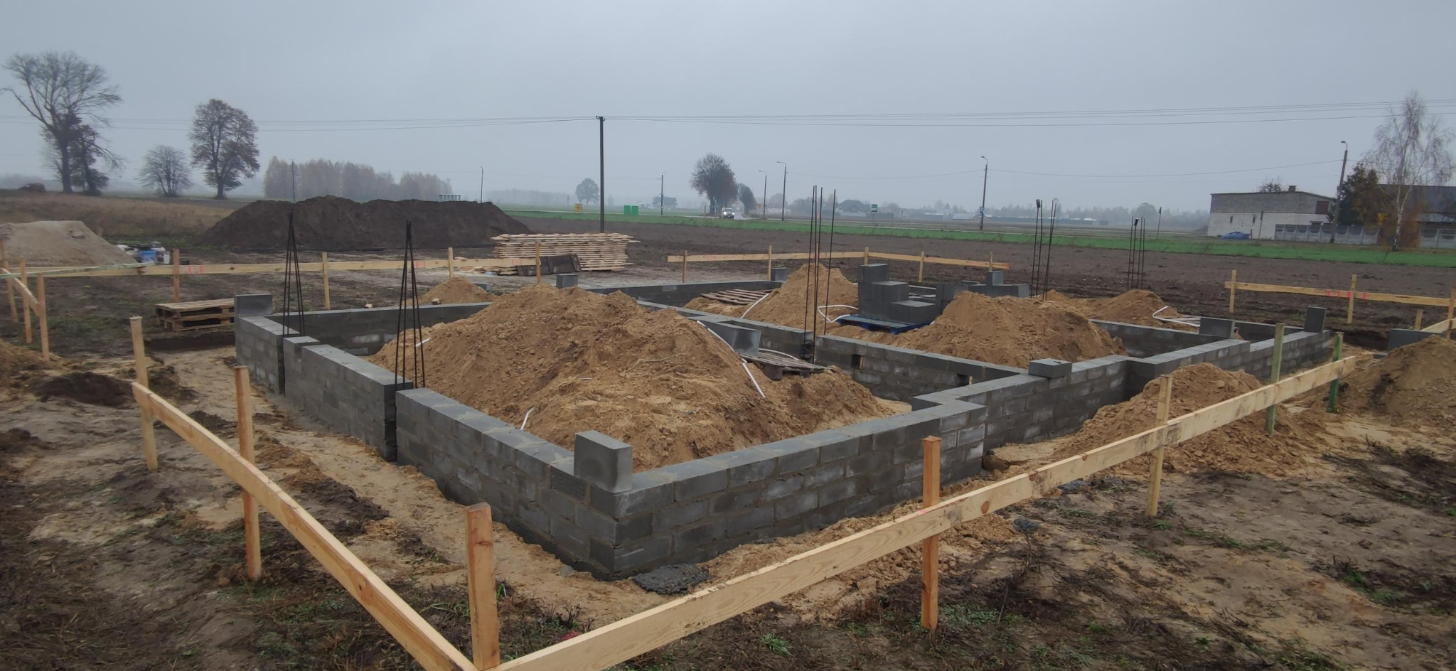 Fundamenty domu jednorodzinnego w trakcie budowy, z widocznymi bloczkami betonowymi, ziemią i drewnianymi elementami szalunku. Pochmurne niebo w tle.