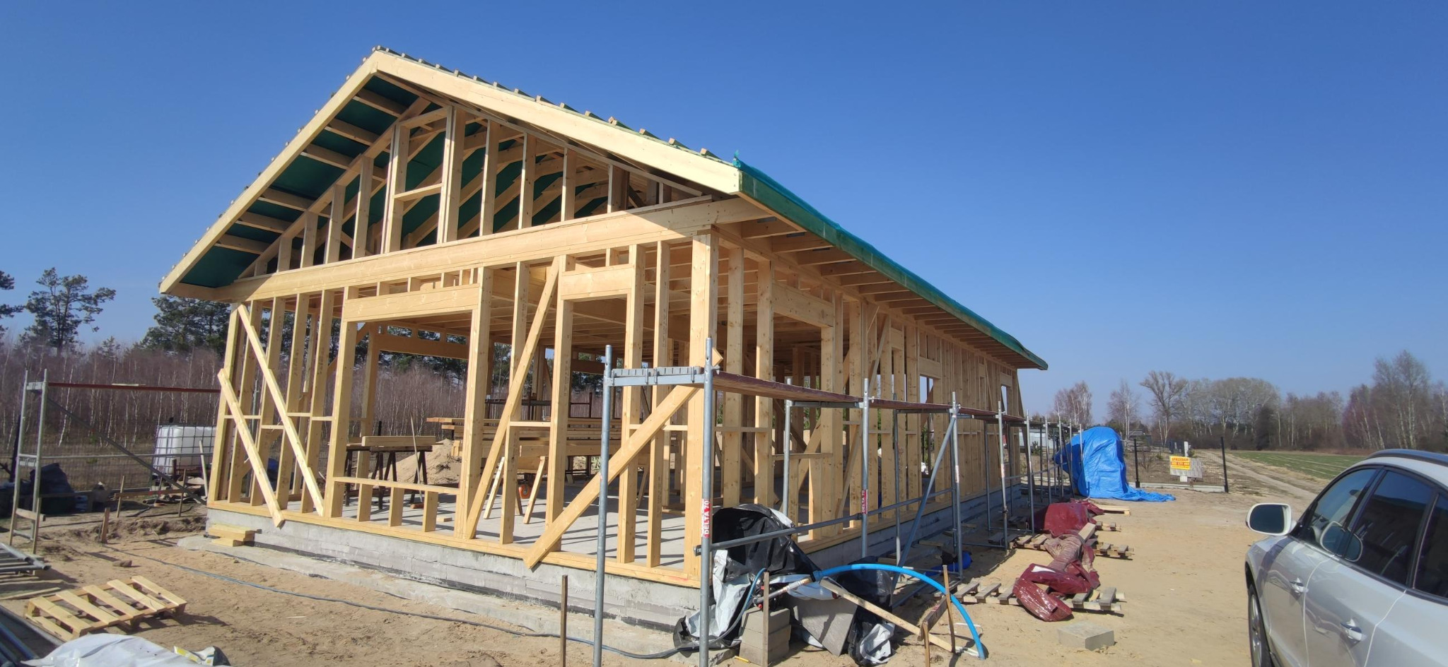 Drewniany szkielet domu jednorodzinnego w trakcie budowy, widok z zewnątrz, słoneczny dzień, widoczne rusztowania i materiały budowlane. Konstrukcja w stanie surowym otwartym.