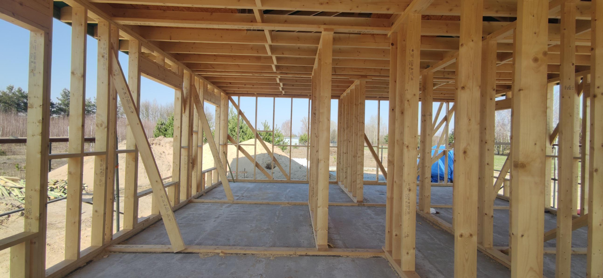 Surowa konstrukcja domu z drewna, widok z wnętrza. Jasne drewno, betonowa podłoga, w tle drzewa i piasek. Widoczne stemple z oznaczeniami na elementach konstrukcyjnych.