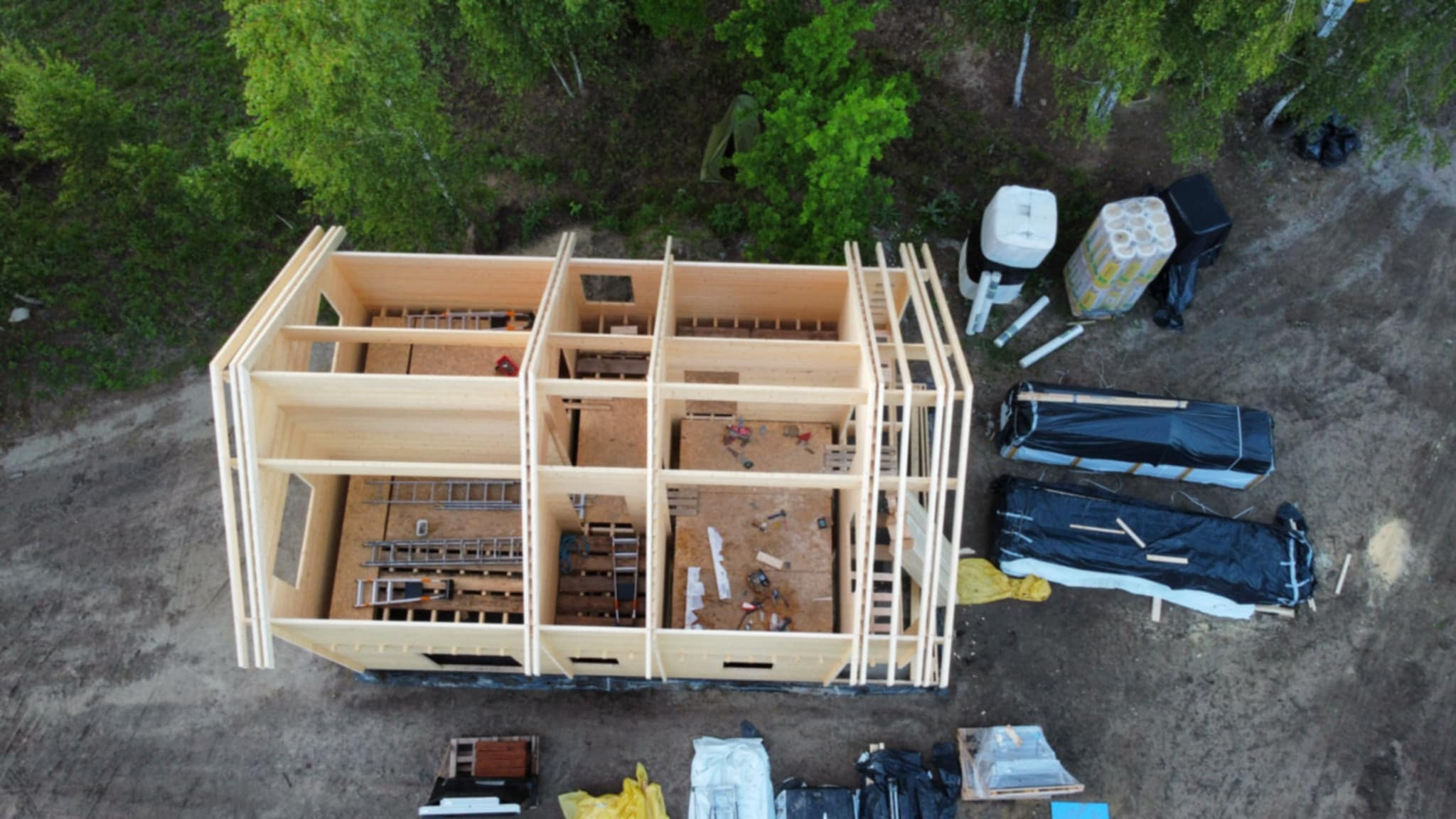 Wznoszenie szkieletu drewnianego domu z widokiem z góry. Widoczne narzędzia i materiały budowlane. Konstrukcja w surowym stanie, otoczona zielenią i ziemią.