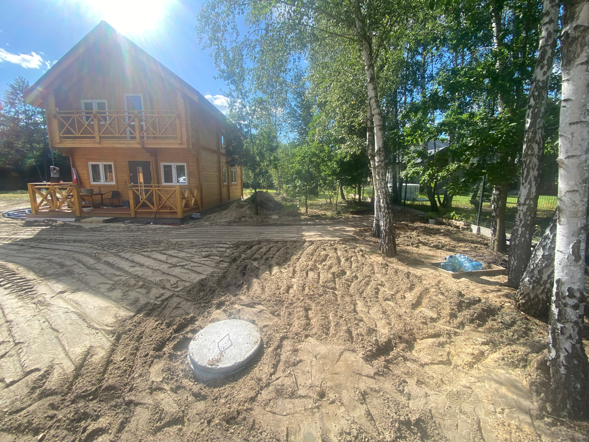 Drewniany dom z balkonem w otoczeniu drzew, widoczny właz kanalizacyjny i przygotowany teren pod budowę. Słoneczny dzień.