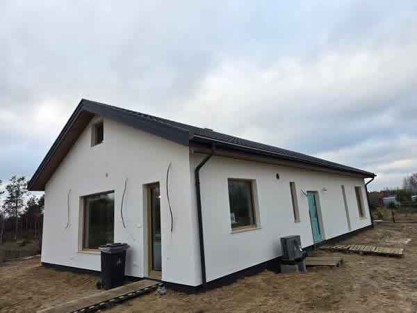 Nowoczesny, parterowy dom jednorodzinny z białym tynkiem i czarnym dachem, widoczny z boku, z elementami wykończeniowymi w trakcie realizacji.