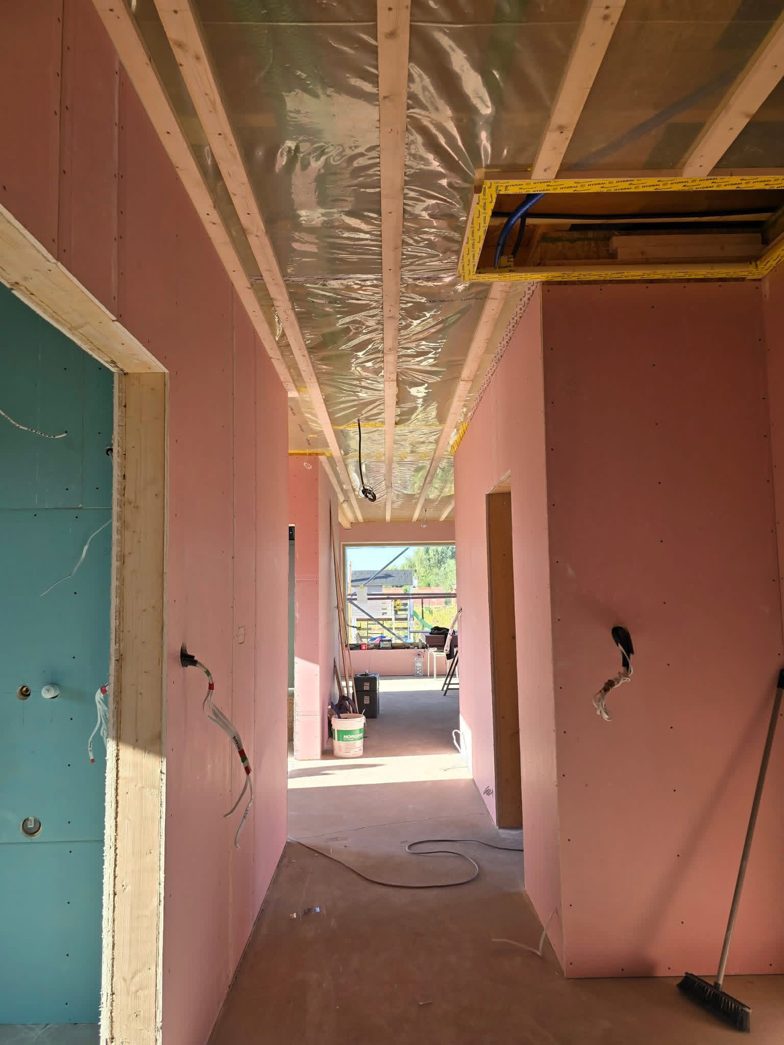 Wnętrze w trakcie wykańczania: różowe ściany z płyt gipsowych, drewniana konstrukcja sufitu z folią, widoczne instalacje elektryczne i otwór rewizyjny. Jasne światło dzienne.