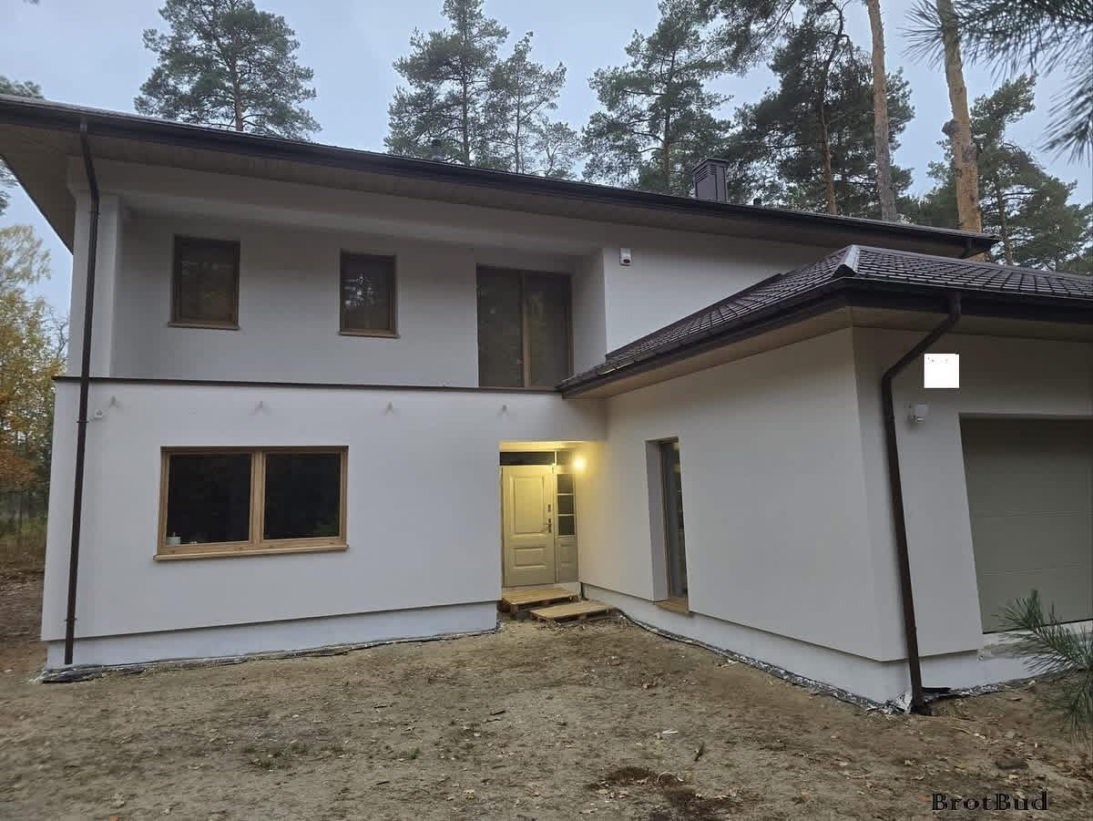 Dwukondygnacyjny dom z garażem w stylu nowoczesnej stodoły, wykończony jasnym tynkiem, z drewnianą stolarką okienną i ciemną dachówką, otoczony drzewami.