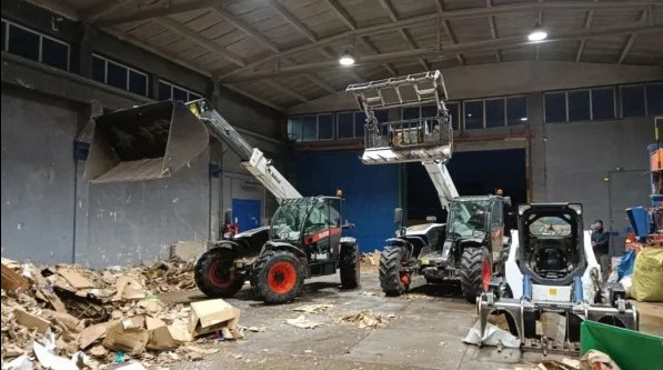 Wnętrze hali z trzema maszynami budowlanymi, w tym dwiema ładowarkami teleskopowymi Bobcat i miniładowarką, oraz stertą pogniecionych kartonów na podłodze.