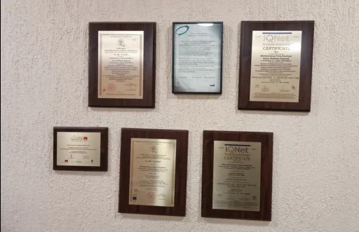 Ściana z certyfikatami i dyplomami w drewnianych ramkach, w tym certyfikaty IQNet, prezentowane na teksturowanej, jasnej ścianie.