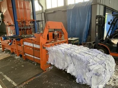 Pomarańczowa prasa hydrauliczna zgniatająca makulaturę w kostkę, widoczny wózek widłowy w tle.