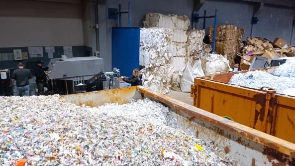 Proces niszczenia dokumentów: pracownicy obsługują przemysłową niszczarkę, a pocięte dokumenty gromadzone są w dużym, metalowym kontenerze. W tle widoczne bele sprasowanego papieru i kartonów.
